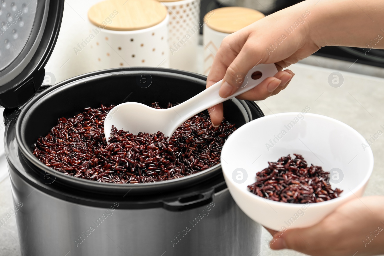 Photo of Woman putting brown rice into bowl from multi cooker in kitchen, closeup