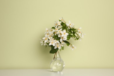 Bouquet of beautiful jasmine flowers in glass vase on table near light green wall