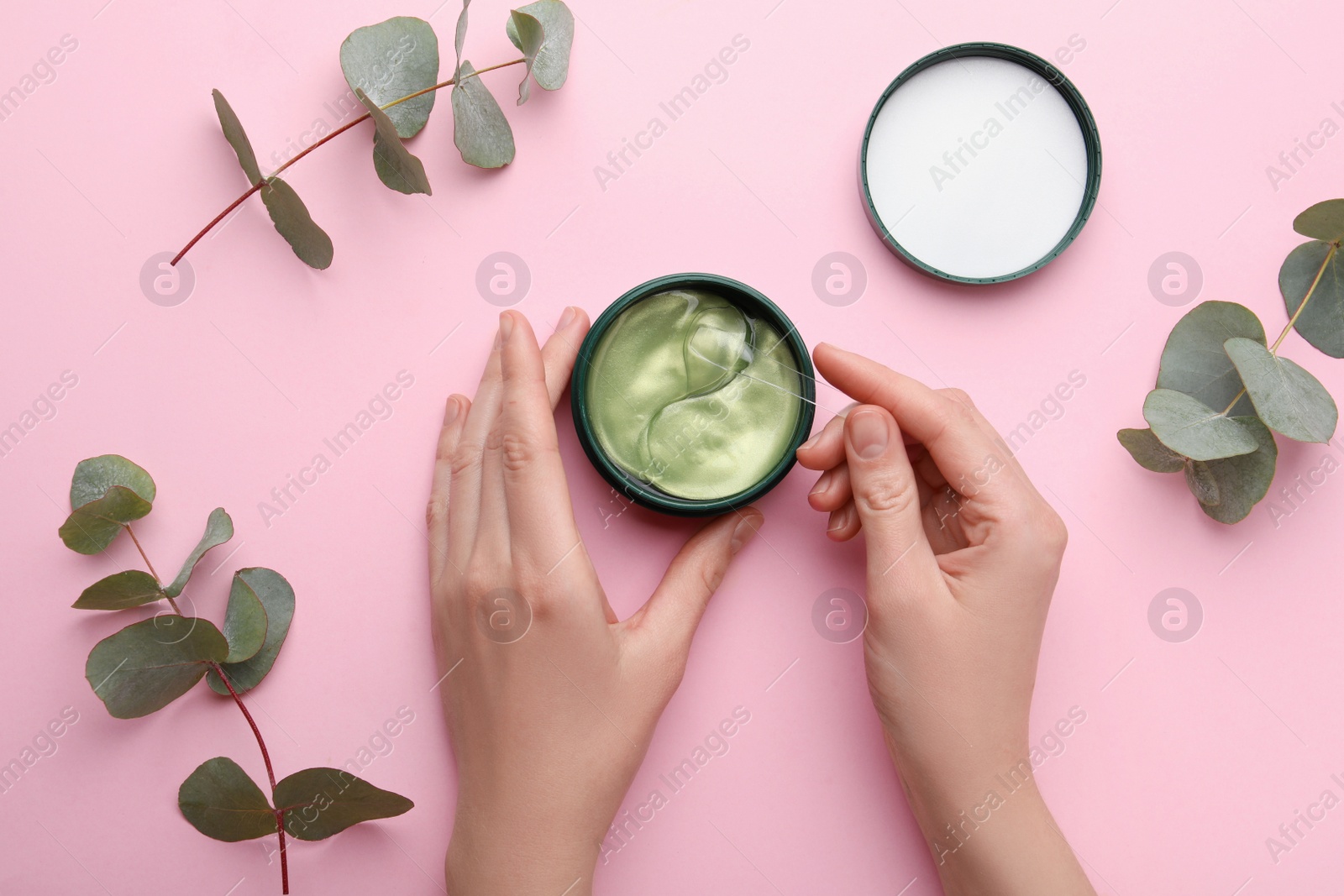Photo of Woman taking under eye patch with spatula out of jar near green twigs on light pink background, top view. Cosmetic product
