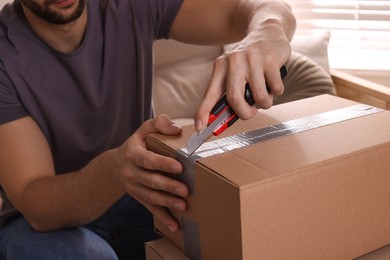 Photo of Man using utility knife to open parcel at home, closeup