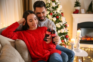 MYKOLAIV, UKRAINE - JANUARY 27, 2021: Young couple holding bottles of Coca-Cola in room decorated for Christmas