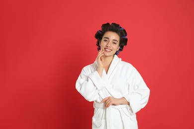 Happy young woman in bathrobe with hair curlers on red background, space for text