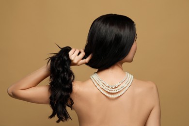 Young woman wearing elegant pearl necklace on brown background, back view