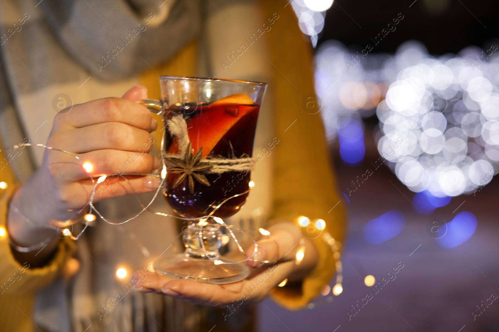 Photo of Woman with glass cup of mulled wine and garland at winter fair, closeup. Space for text