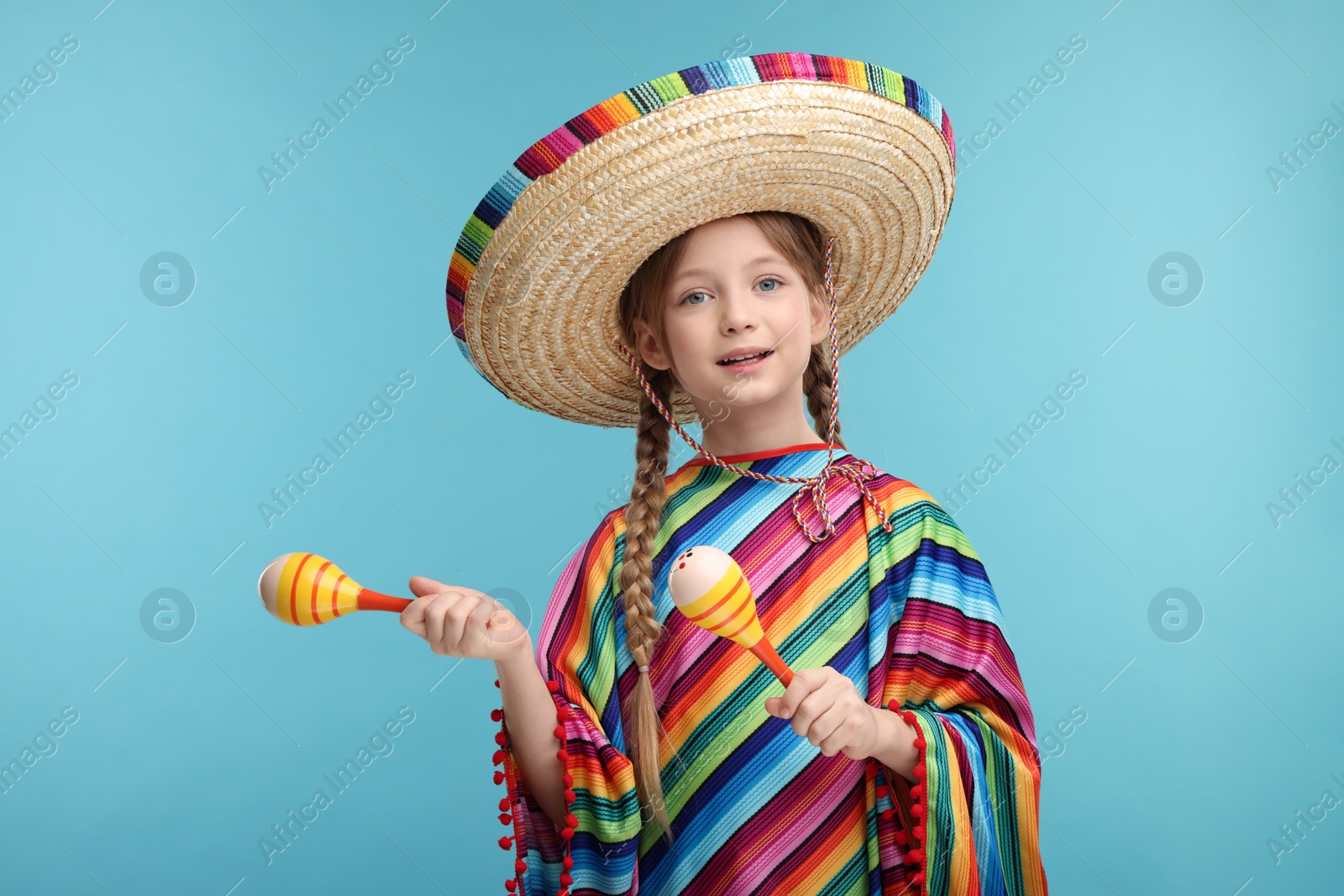 Photo of Cute girl in Mexican sombrero hat and poncho dancing with maracas on light blue background