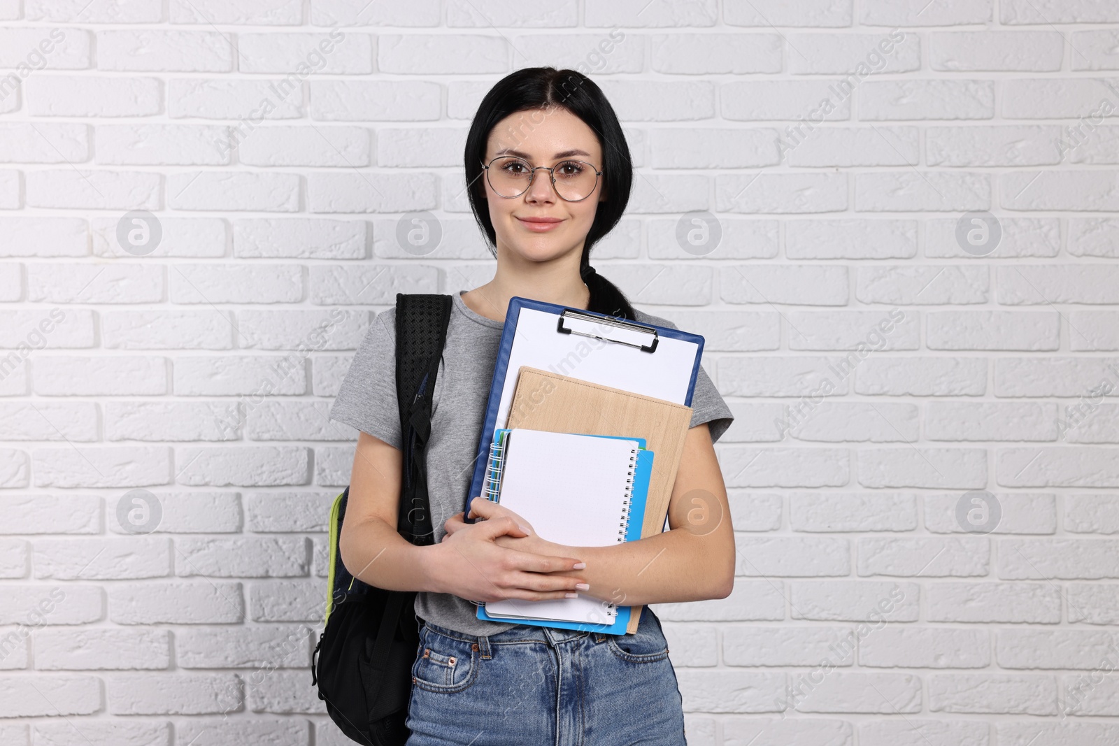 Photo of Student with notebooks, clipboard and backpack near white brick wall