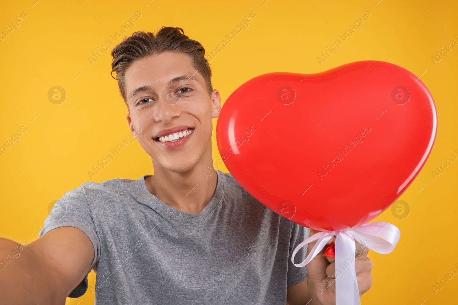 Photo of Happy man holding red heart shaped balloon and taking selfie on orange background