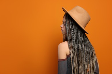 Beautiful woman with long african braids and hat on orange background, space for text