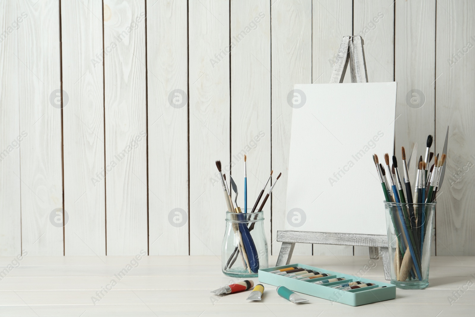 Photo of Easel with blank canvas, paints and brushes on white wooden table. Space for text