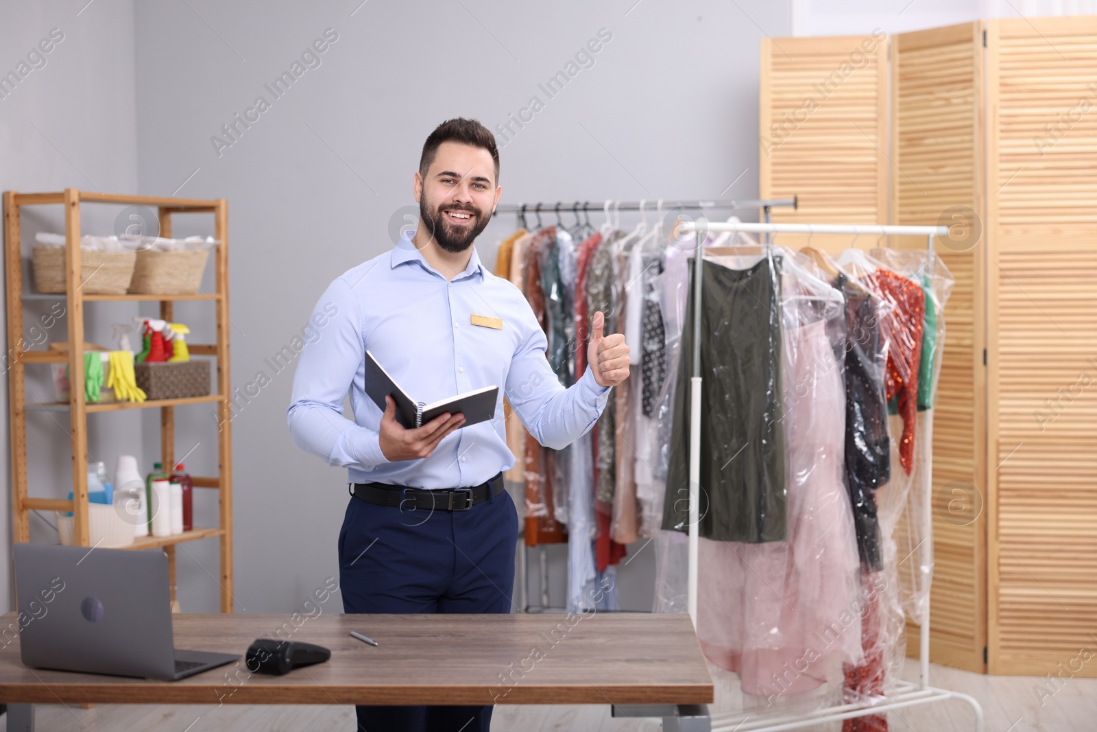 Photo of Dry-cleaning service. Happy worker with notebook showing thumb up in workplace indoors, space for text