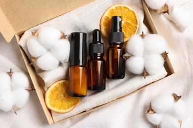 Photo of Box with bottles of organic cosmetic products, dried orange slices and cotton flowers on white fabric, flat lay