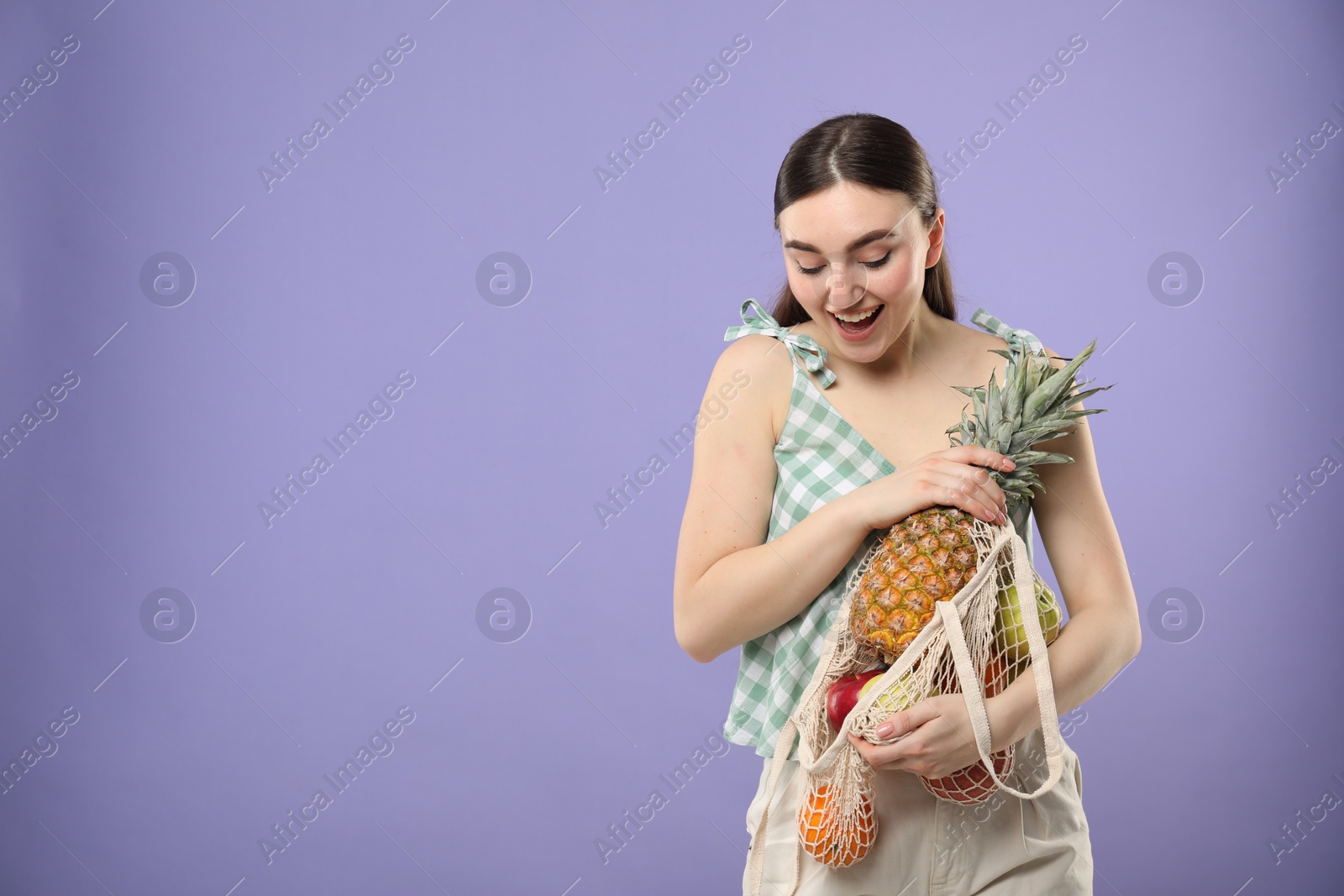 Photo of Woman with string bag of fresh fruits on violet background, space for text