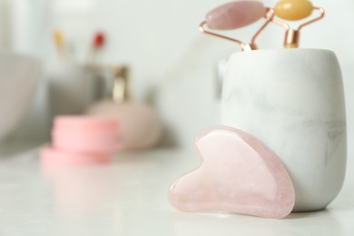 Photo of Rose quartz gua sha tool near holder with natural face rollers on white countertop in bathroom, closeup. Space for text