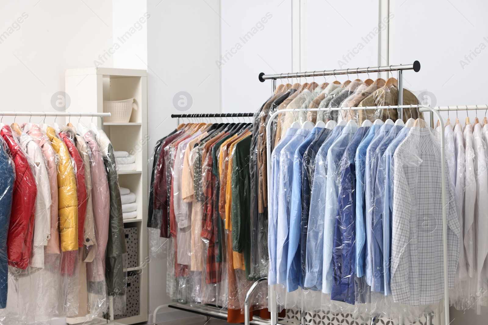 Photo of Dry-cleaning service. Many different clothes in plastic bags hanging from racks indoors