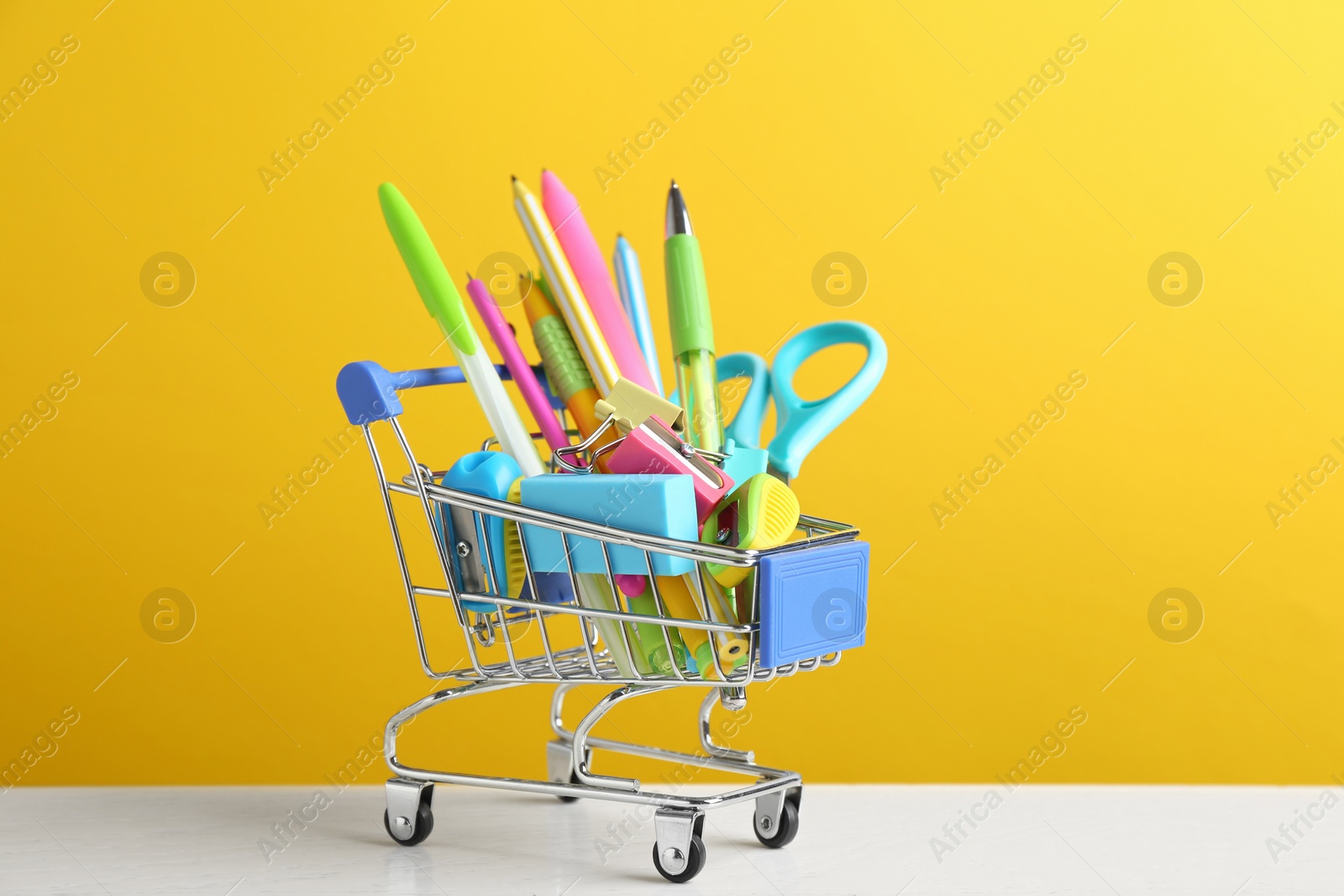 Photo of Small shopping cart with different school stationery on white wooden table against yellow background