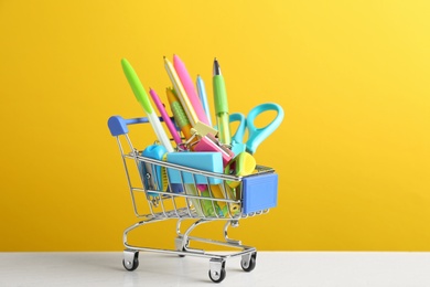 Photo of Small shopping cart with different school stationery on white wooden table against yellow background