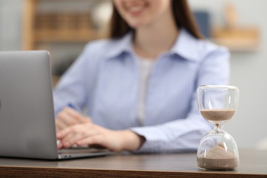 Photo of Hourglass with flowing sand on desk. Woman using laptop indoors, selective focus