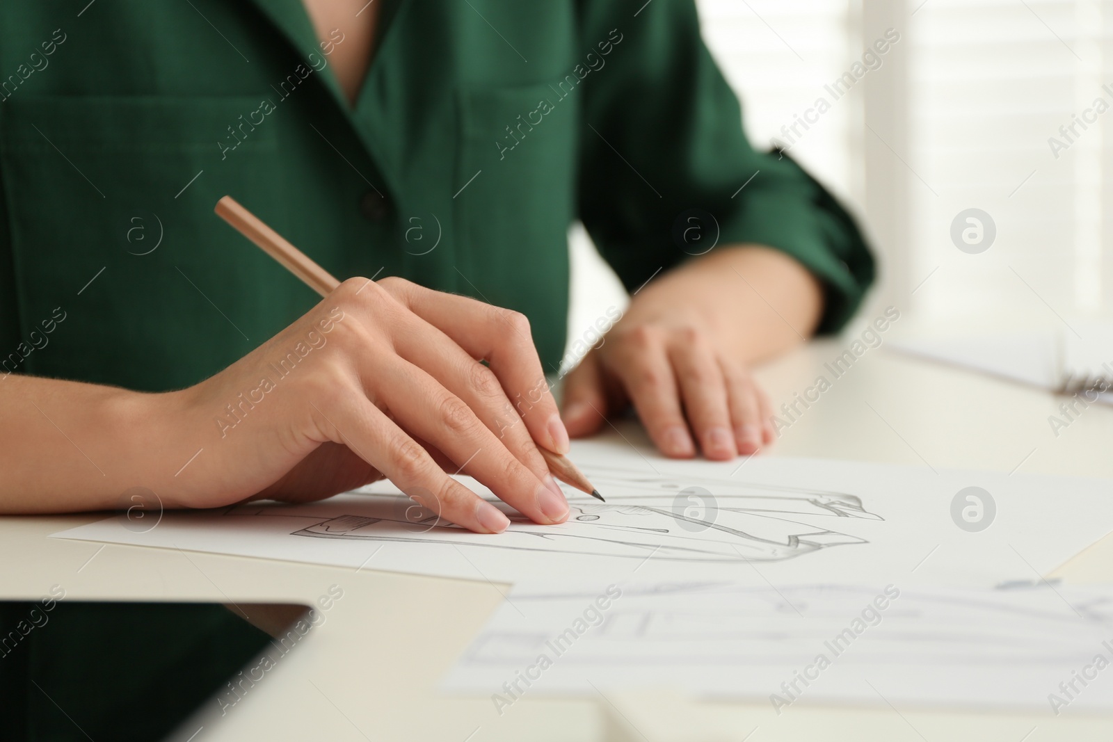 Photo of Woman drawing clothes with pencil on sheet of paper at white table, closeup