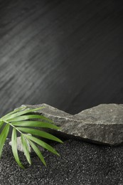 Photo of Presentation of product. Stone podium and palm leaves on black sand against dark textured background. Space for text