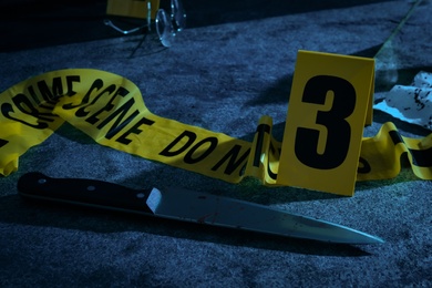 Photo of Yellow tape, crime scene marker and bloody knife on grey stone table at night
