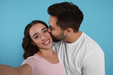 Photo of Man kissing his smiling girlfriend while she taking selfie on light blue background