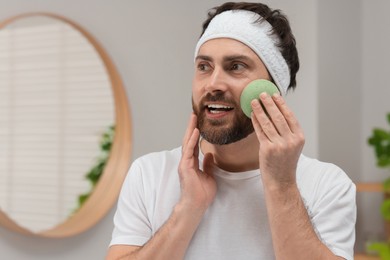 Man with headband washing his face using sponge in bathroom, space for text