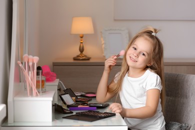 Adorable little girl applying makeup at dressing table indoors
