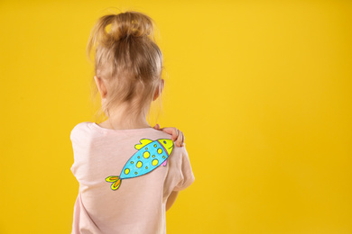 Photo of Little girl with paper fish on back against yellow background, space for text. April fool's day