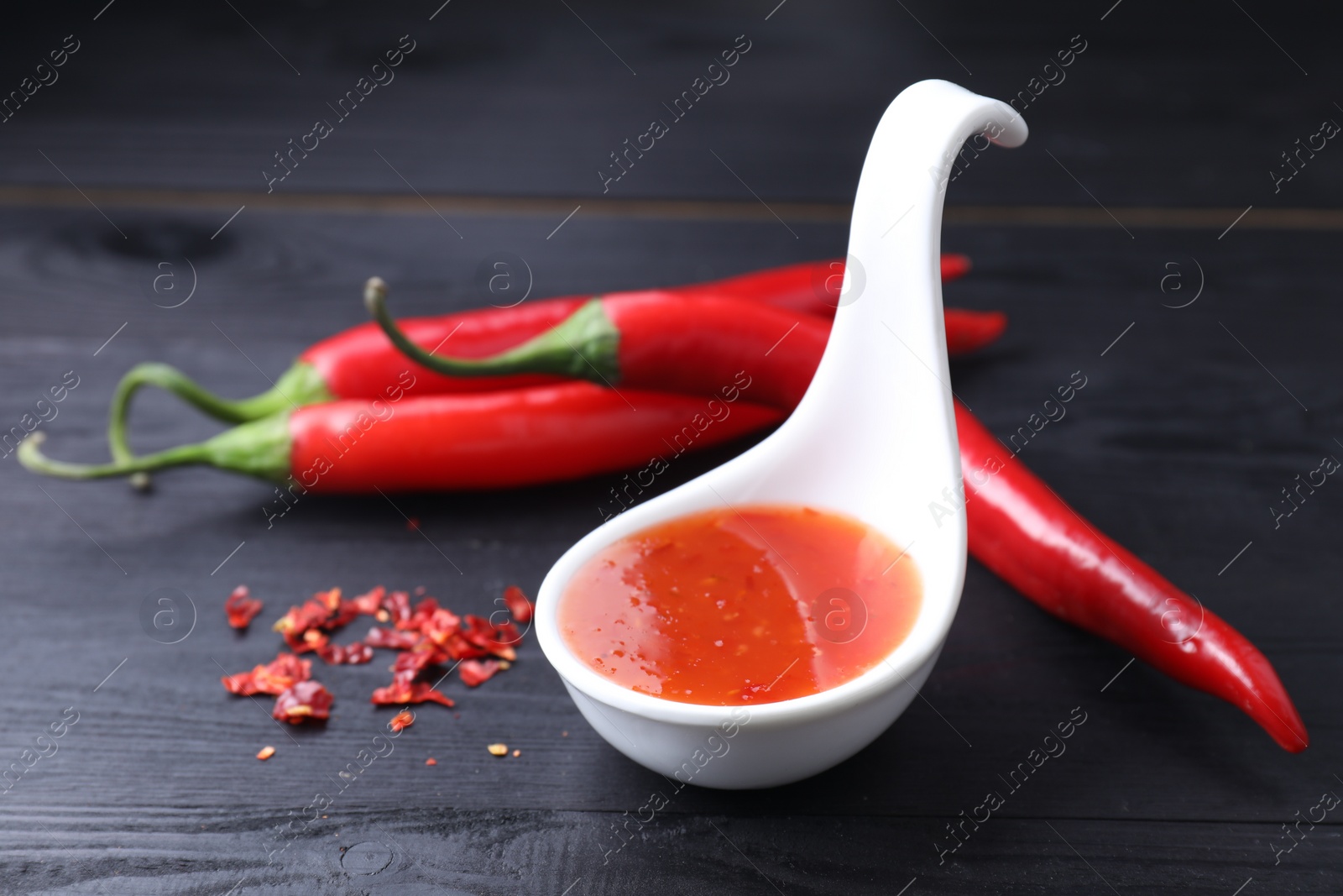 Photo of Spicy chili sauce in spoon and peppers on black wooden table, closeup