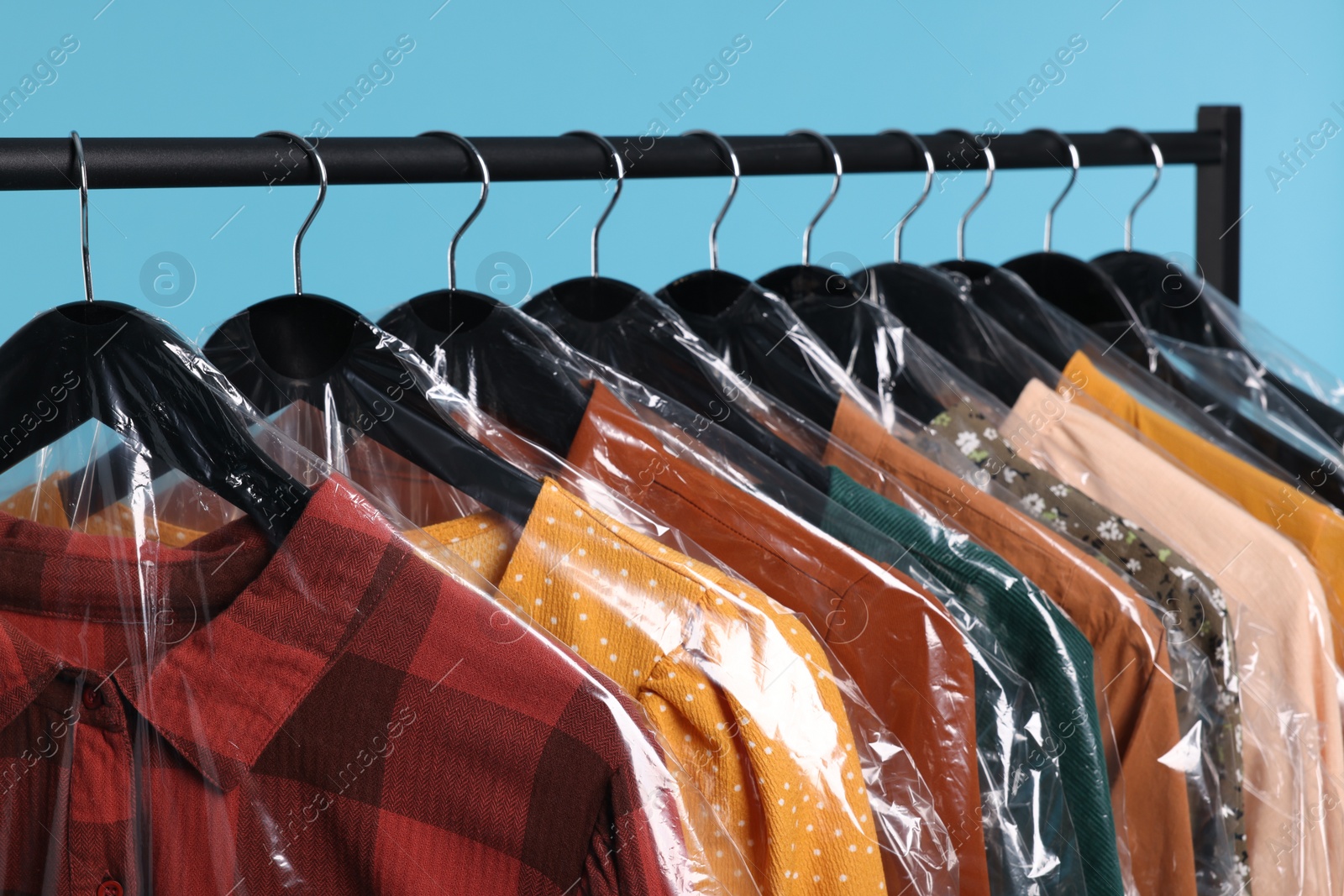 Photo of Dry-cleaning service. Many different clothes in plastic bags hanging on rack against light blue background, closeup