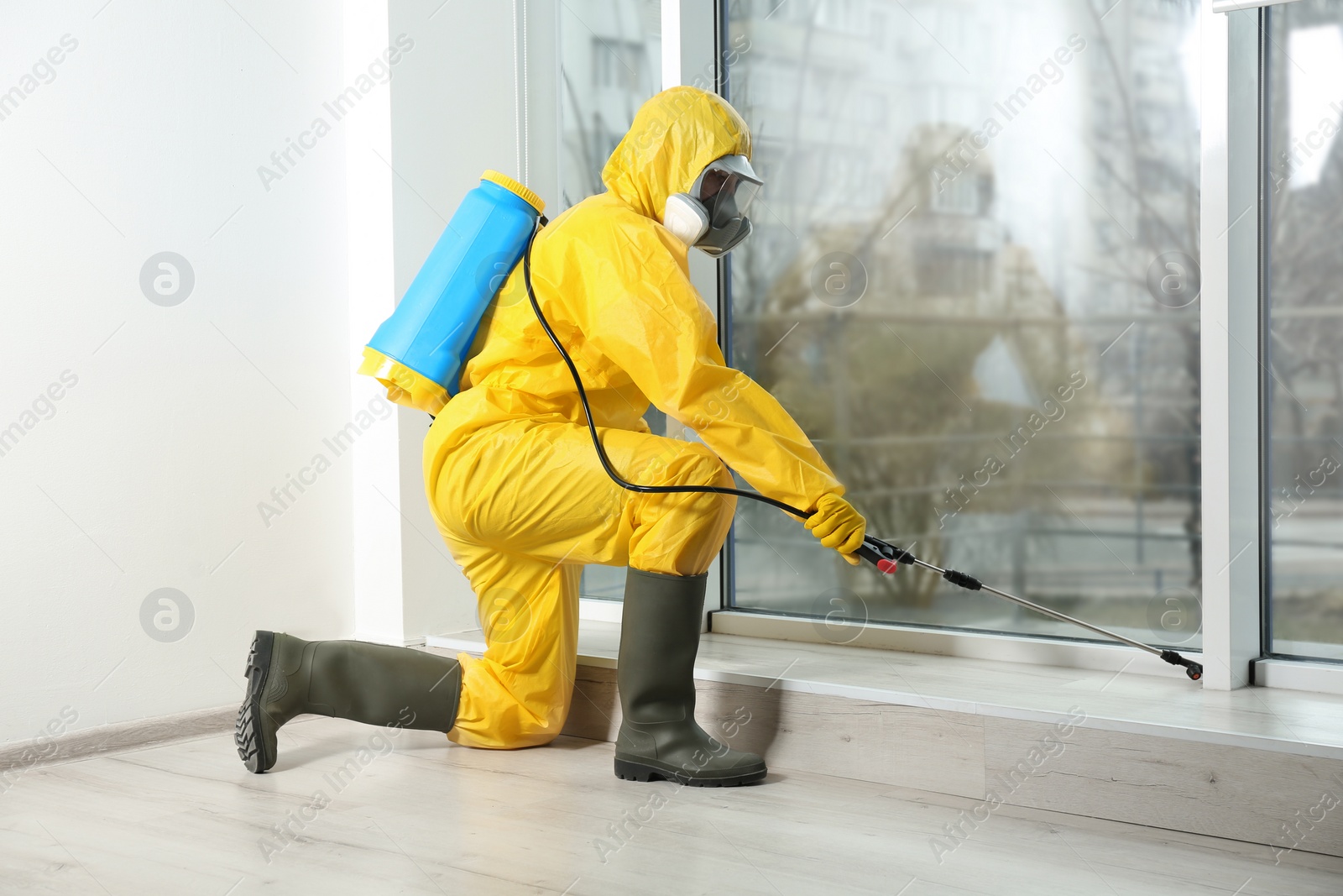 Photo of Pest control worker in protective suit spraying pesticide near window indoors