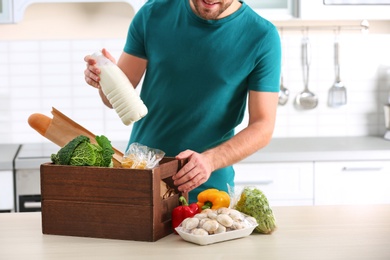 Photo of Man with wooden crate full of products at table in kitchen. Food delivery service