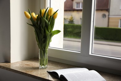 Bouquet of beautiful yellow tulip flowers in glass vase and open book on windowsill. Space for text
