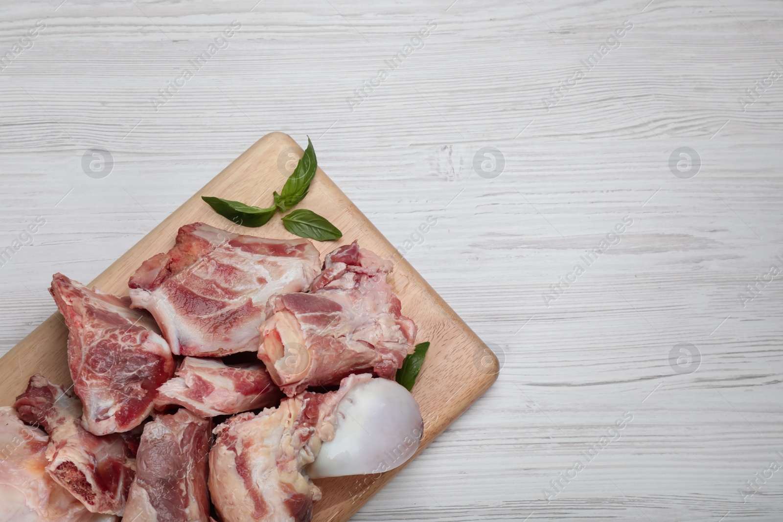 Photo of Cutting board with raw chopped meaty bones and basil on white wooden table, top view. Space for text