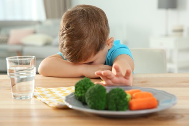 Unhappy little boy refusing to eat vegetables at table in room