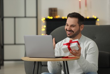 Celebrating Christmas online with exchanged by mail presents. Happy man with gift box during video call on laptop at home