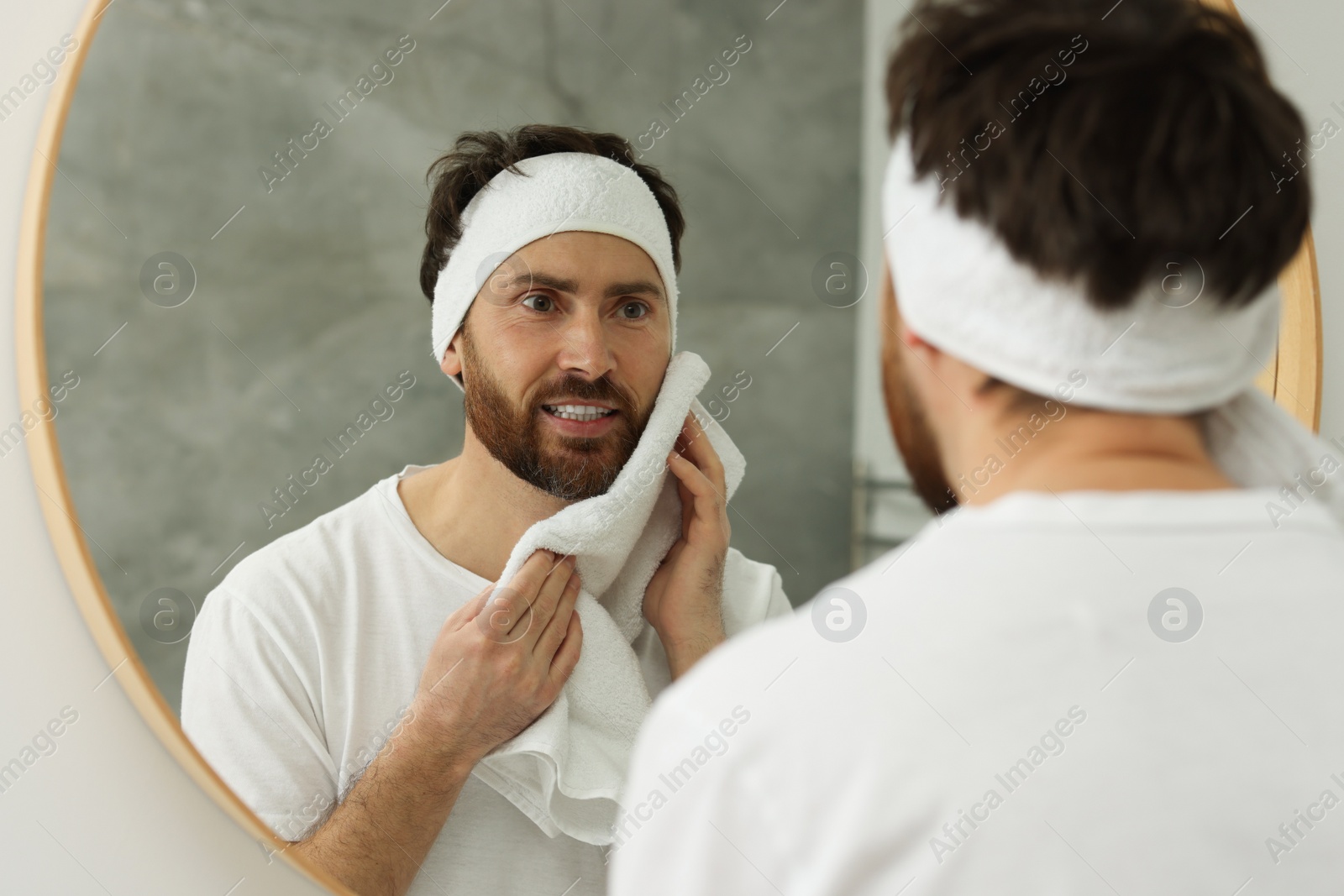 Photo of Washing face. Man with headband and towel near mirror in bathroom