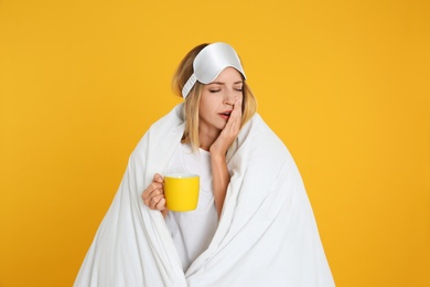 Young woman in sleeping mask wrapped with blanket holding cup on yellow background