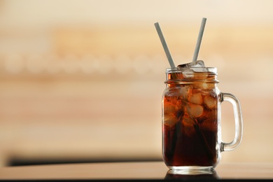 Photo of Mason jar of cola with ice on table against blurred background. Space for text