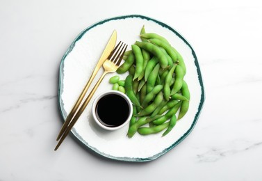Photo of Green edamame beans in pods served with soy sauce on white marble table, top view