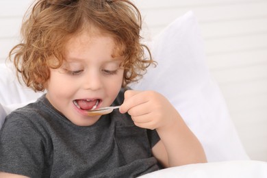 Photo of Cute boy taking measuring spoon with cough syrup on bed. Effective medicine