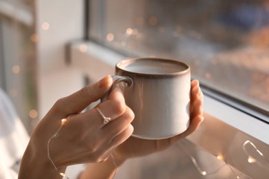 Young woman with cup of hot coffee near window, closeup