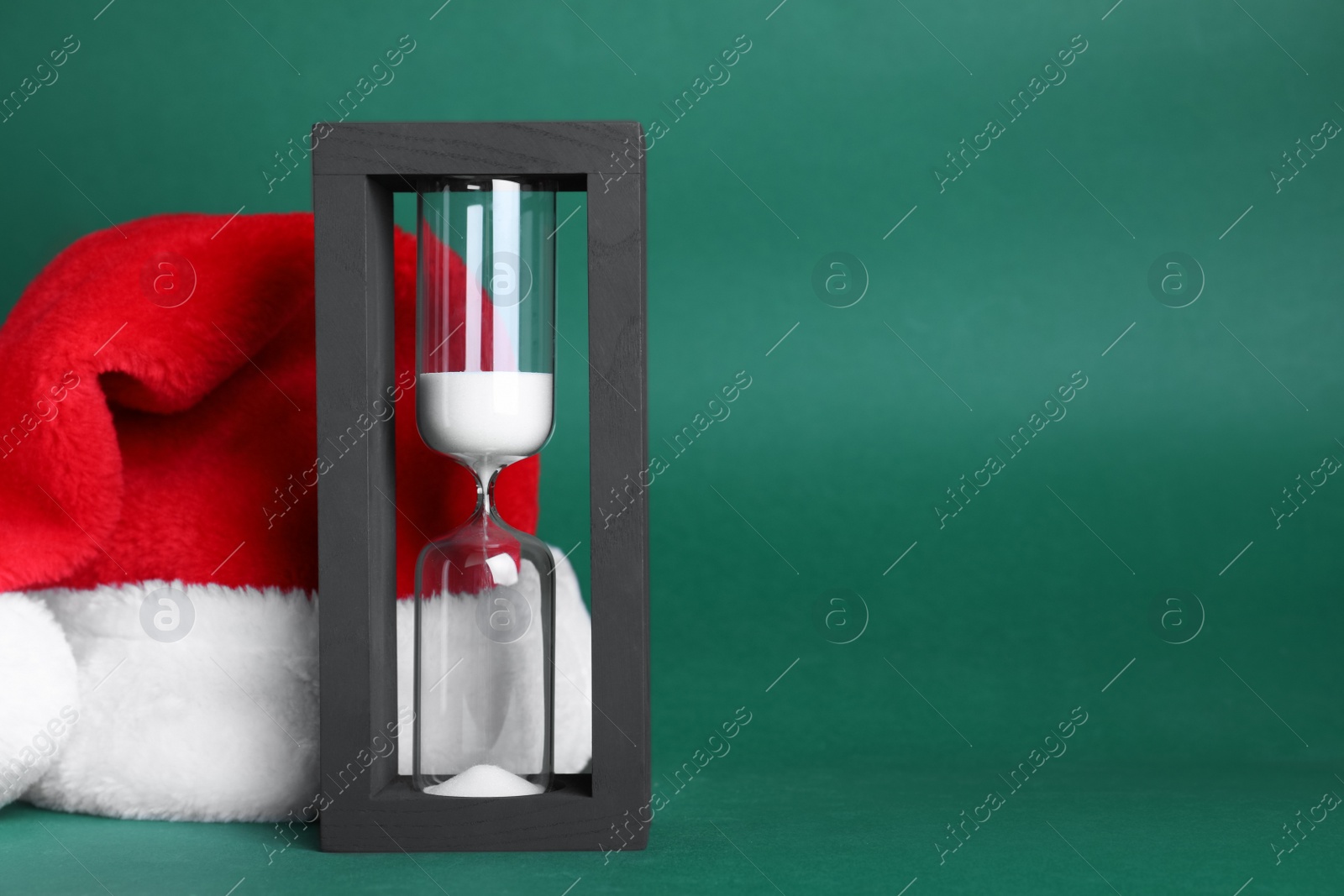 Photo of Hourglass and Santa hat on green background, space for text. Christmas countdown