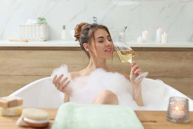 Photo of Beautiful woman with glass of wine enjoying bubble bath at home