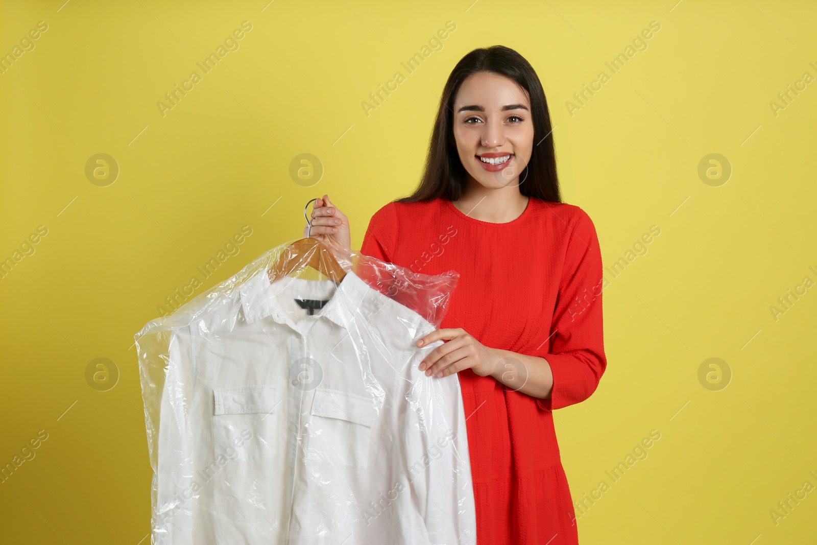 Photo of Young woman holding hanger with shirt in plastic bag on yellow background. Dry-cleaning service