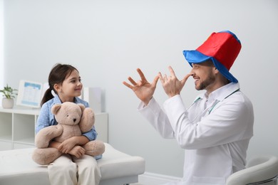 Photo of Pediatrician in funny hat playing with little girl during visit at hospital