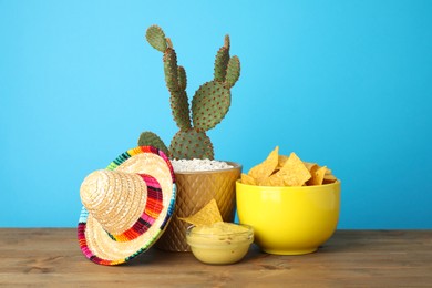 Photo of Mexican sombrero hat, cactus, nachos chips and guacamole in bowls on wooden table against light blue background