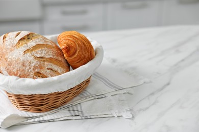 Wicker bread basket with freshly baked loaf and croissant on white marble table in kitchen, space for text
