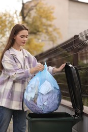 Woman putting garbage bag into recycling bin outdoors