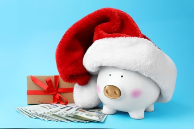 Photo of Piggy bank with Santa hat, dollar banknotes and gift box on light blue background